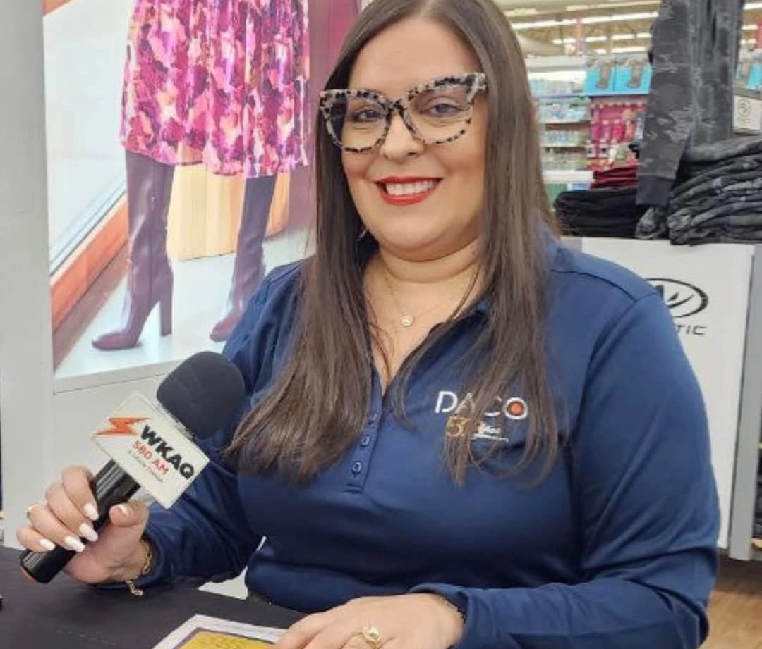 Lisoannette González Ruiz, entonces secretaria del DACO, sonríe mientras sostiene un micrófono de la emisora WKAQ 580 AM durante una actividad pública. Viste una camisa azul marino con el logo del DACO y usa espejuelos de montura tipo ojo de gato con diseño de leopardo.
