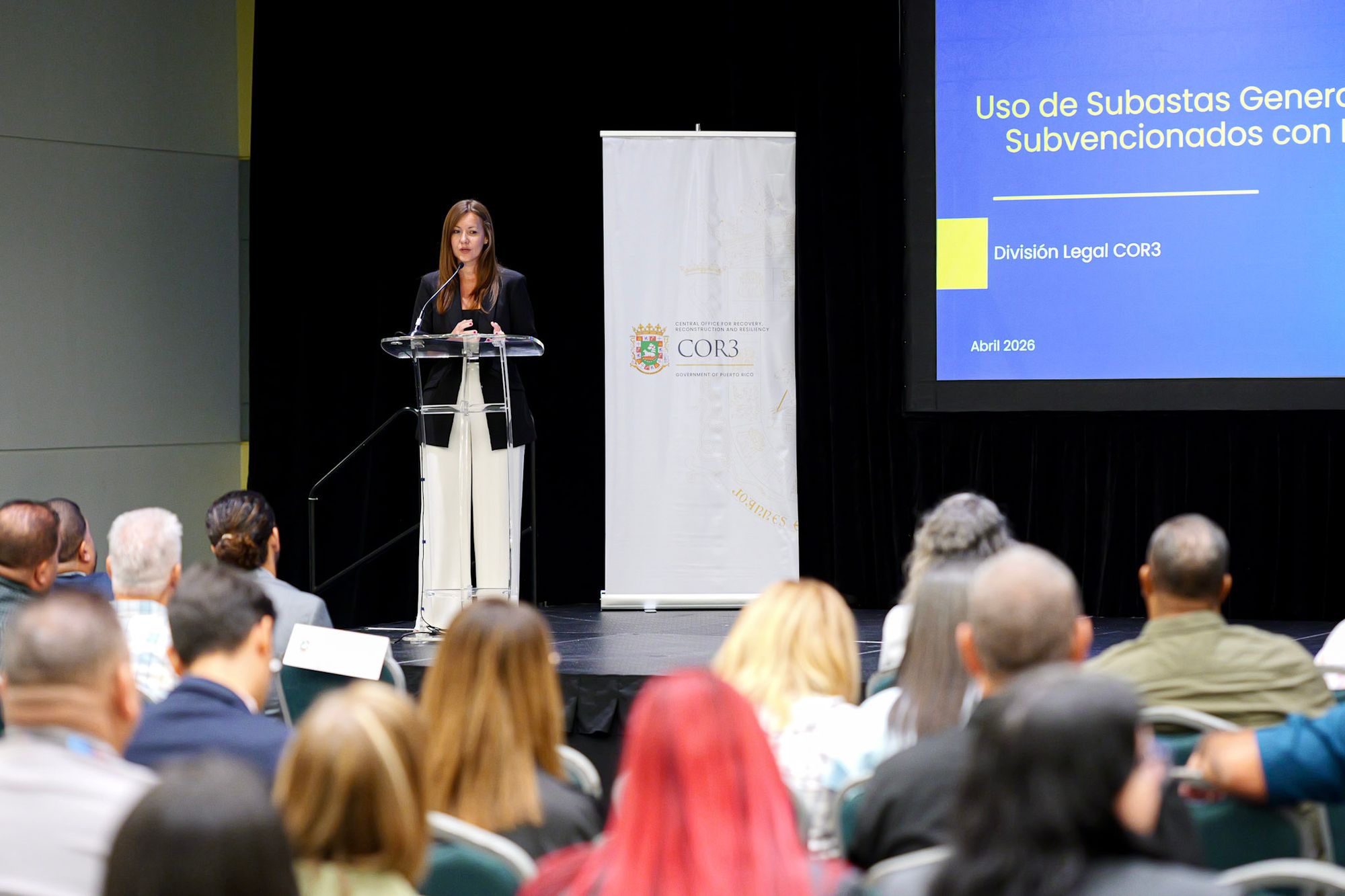 María L. Marín Colón habla desde un podio de acrílico en un salón de conferencias, junto a un banner blanco con el logo de COR3 y frente a una audiencia de funcionarios sentados de espaldas a la cámara, con una pantalla azul al fondo que proyecta una presentación sobre el uso de subastas en proyectos subvencionados con fondos federales.