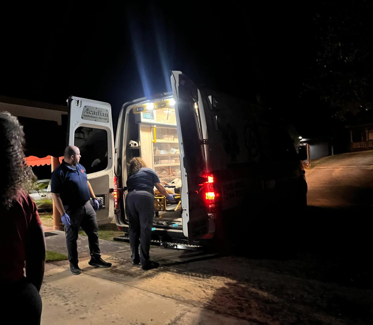Escena nocturna de una ambulancia con las puertas traseras abiertas e iluminada por dentro, mientras dos personas con guantes atienden una camilla en la parte posterior del vehículo, estacionado frente a una residencia.