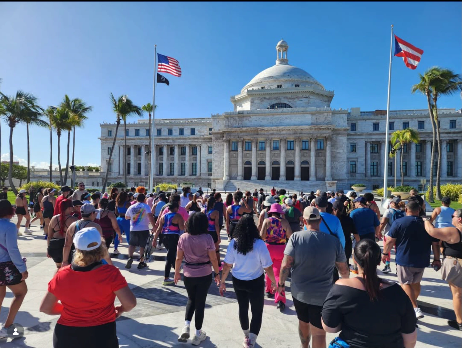 Un grupo numeroso de personas con ropa deportiva, vistas de espaldas, hace ejercicios de pie sobre la explanada de mármol frente al Capitolio de Puerto Rico, con su cúpula blanca al fondo bajo un cielo despejado. A la izquierda ondean la bandera de Estados Unidos y la de los prisioneros de guerra, a la derecha la bandera de Puerto Rico, y palmas reales bordean el edificio.